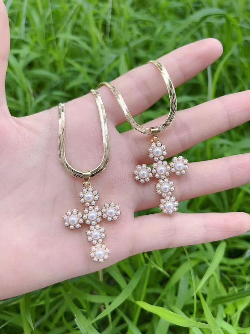 Gold cross-shaped earrings with pearl details held in a hand against a grassy background