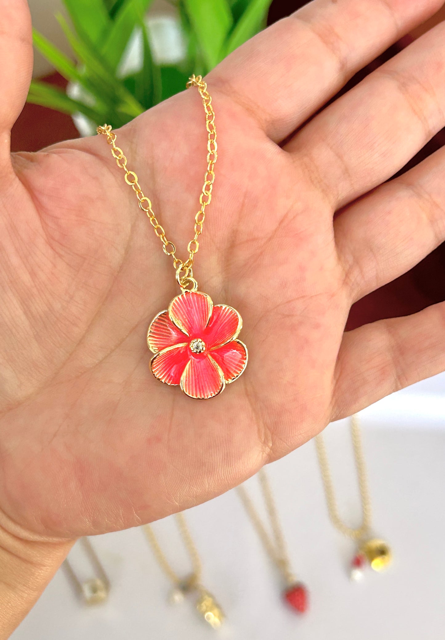 Hand holding a gold necklace with a pink flower pendant against a blurred background