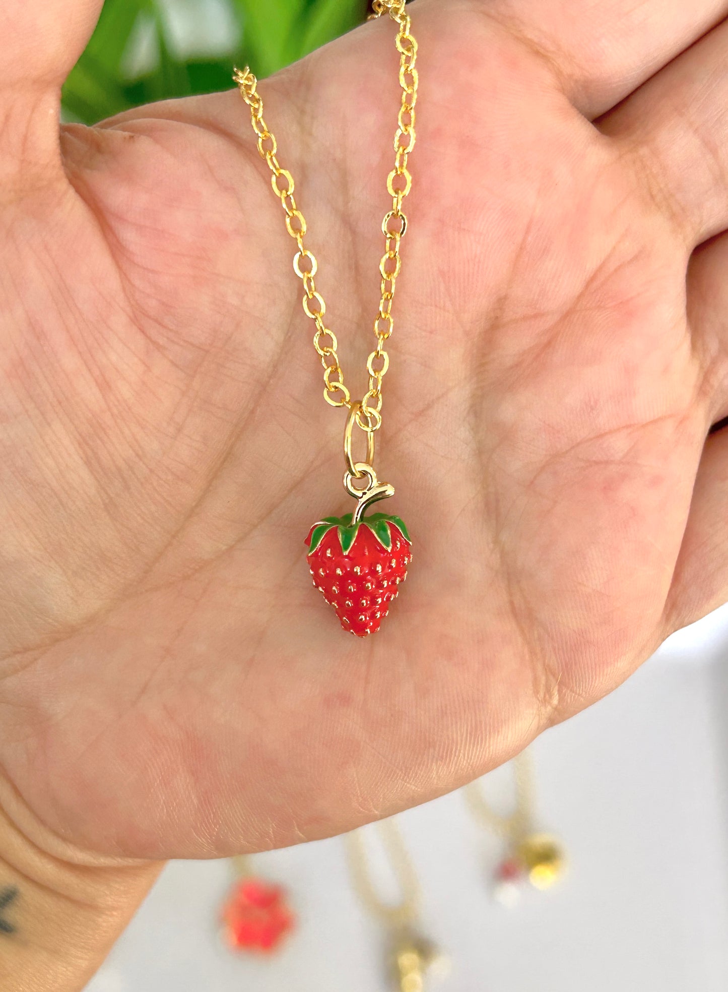 Gold necklace with a strawberry pendant held in a hand against a blurred background