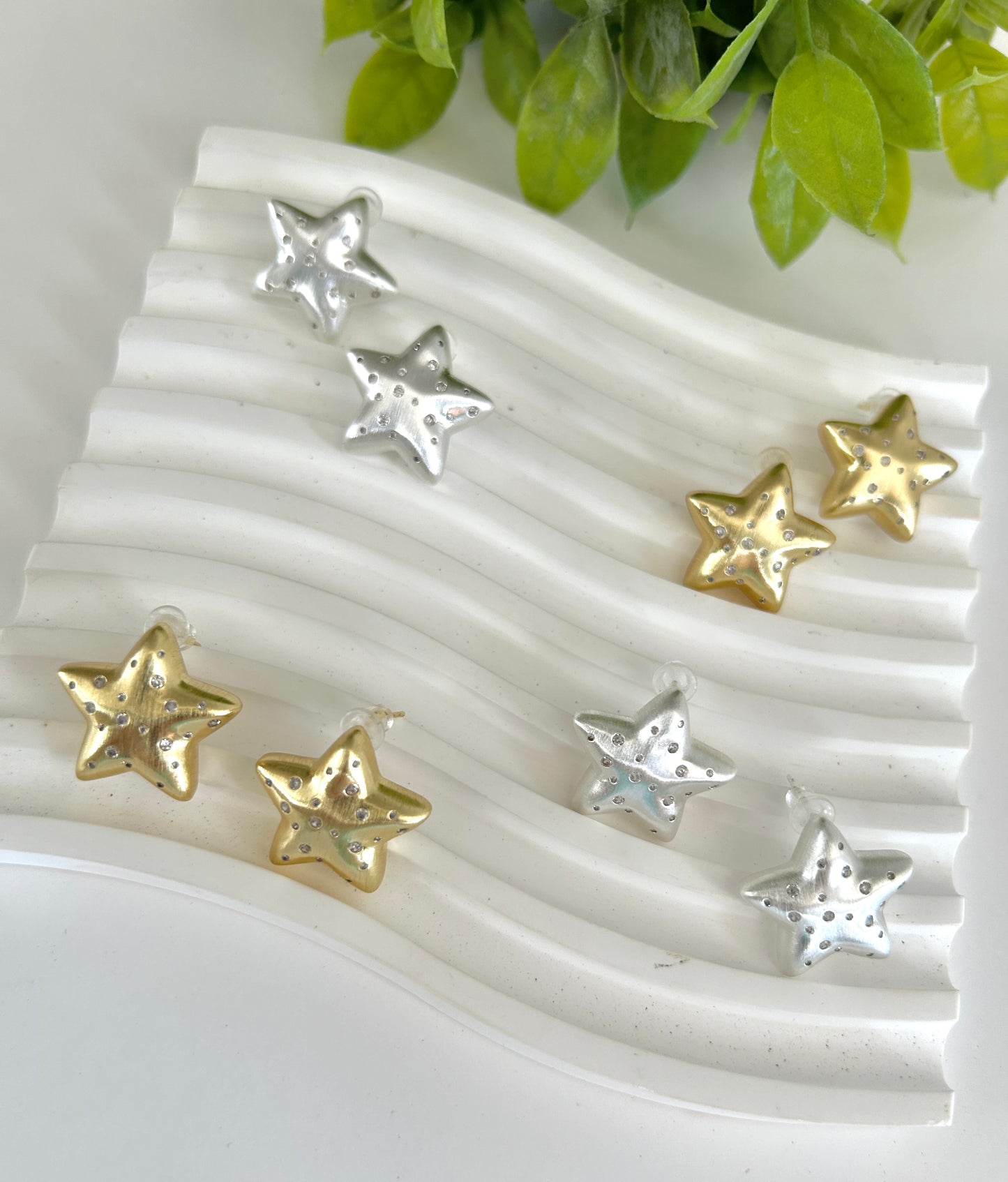 Star-shaped earrings in gold and silver on a textured white surface with green leaves in the background.