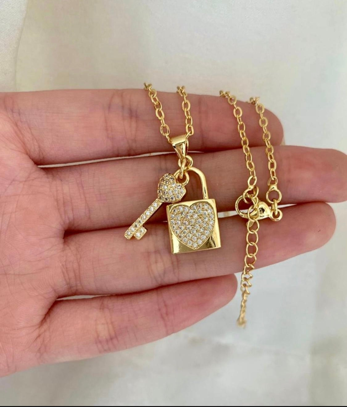 Gold necklace with heart padlock and key pendant held in a hand against a neutral background