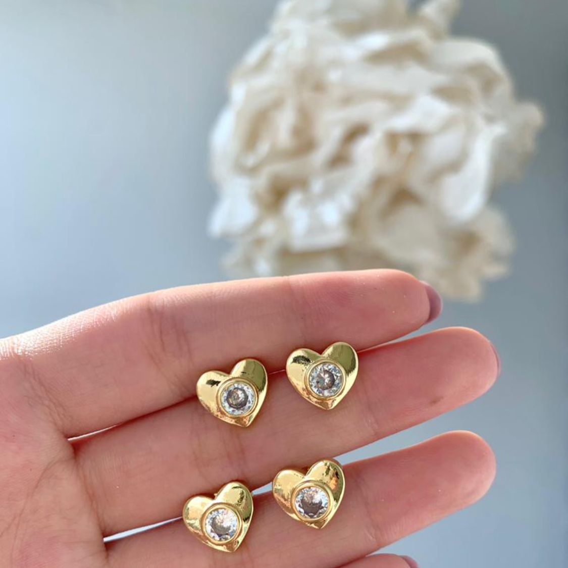 Gold heart-shaped earrings with clear stones held in a hand against a light background