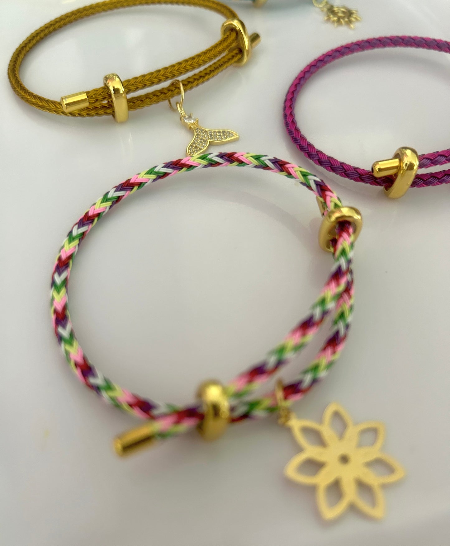 Three colorful braided bracelets with gold charms on a light gray background
