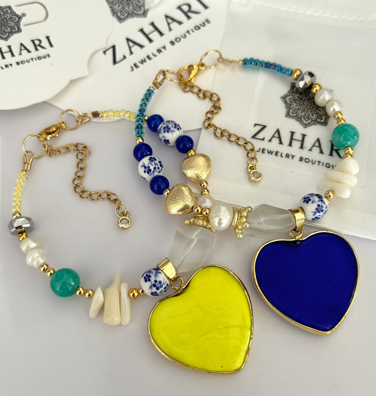 Two beaded bracelets with heart charms on a white surface with 'Zahari Jewelry Boutique' branding.