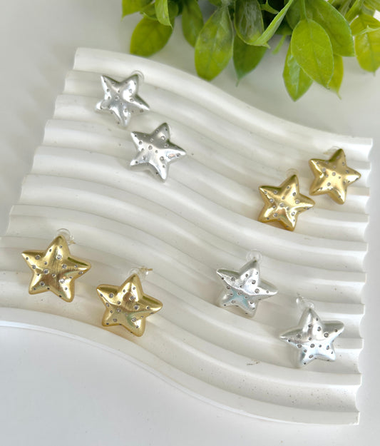 Star-shaped earrings in gold and silver on a textured white surface with green leaves in the background.