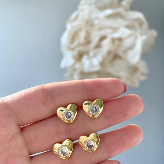 Gold heart-shaped earrings with clear stones held in a hand against a light background