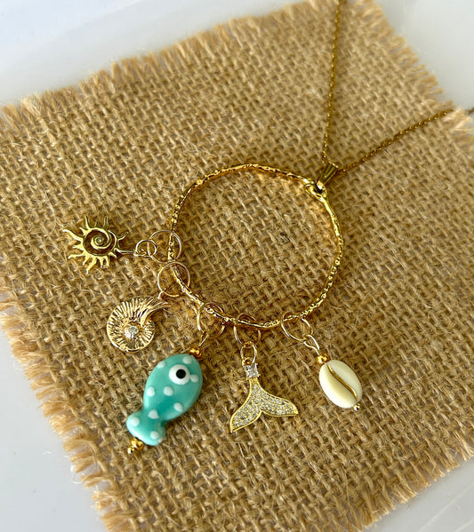 Gold bracelet with various charms including a fish, shell, and mermaid tail on burlap fabric.