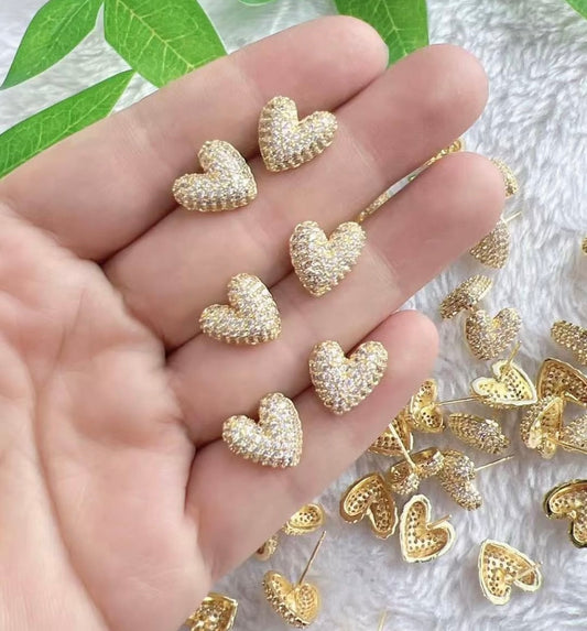 Gold heart-shaped earrings held in a hand with more earrings on a textured surface.