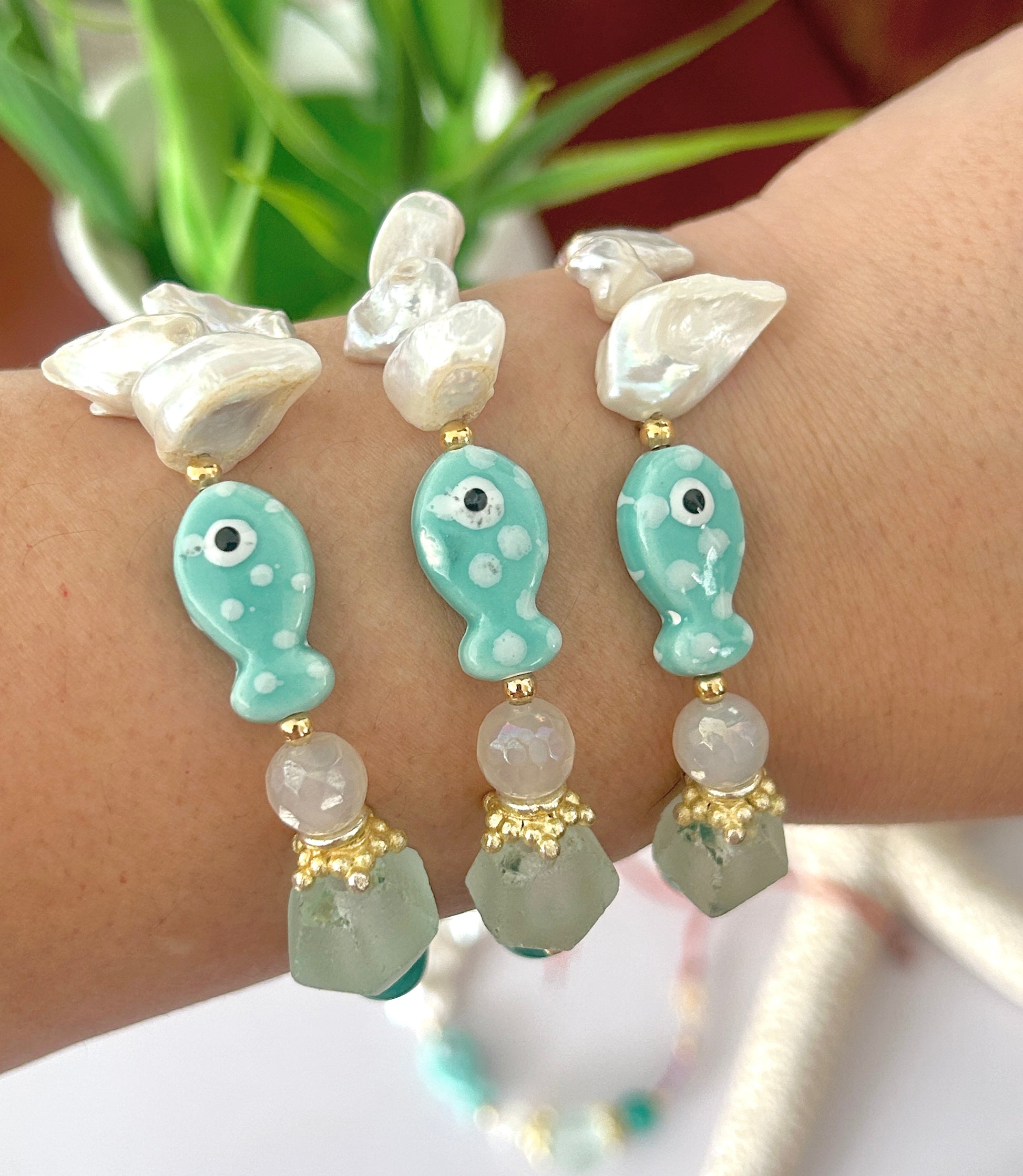 Bracelet with turquoise fish-shaped beads and white pearls on a wrist, with a blurred plant in the background.