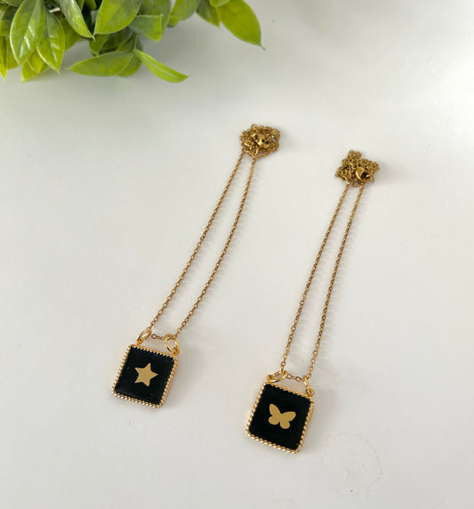 Pair of gold chain earrings with black square pendants featuring star and butterfly designs on a white background.